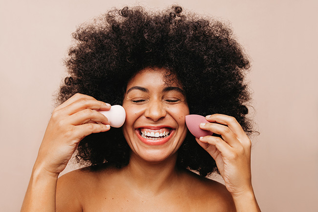 vrouw met krullend haar die lacht en 2 beautyblenders tegen haar gezicht houdt